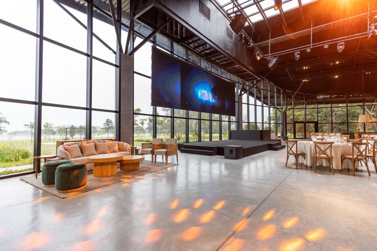 Veranstaltungshalle mit großen Glasfronten, in der eine LED-Wand über einer niedrigen Bühne hängt; davor Lounge-Möbel und eingedeckte Tische