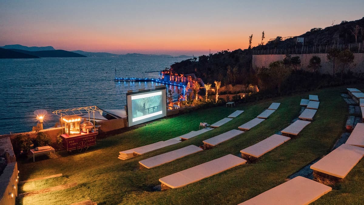 Freiluft-Projektion in der Abenddämmerung: Leinwand am Wasser, davor Sitzreihen auf einer Wiese